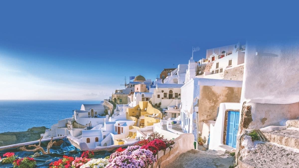 A stunning view of Oia in Santorini, Greece, featuring whitewashed buildings with blue doors and domes, colorful flowers, and the deep blue Aegean Sea stretching into the horizon under a clear sky.
