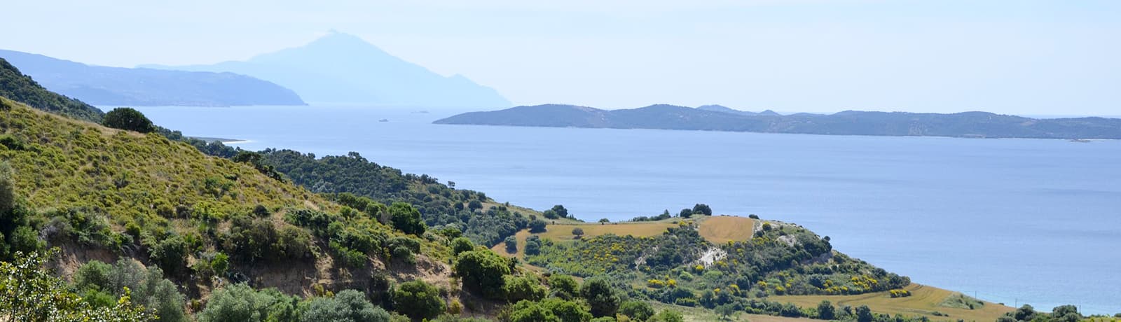 A serene view of the Halkidiki coastline in northern Greece, featuring rolling green hills and blue sea with the distant outline of Mount Athos under a clear sky.