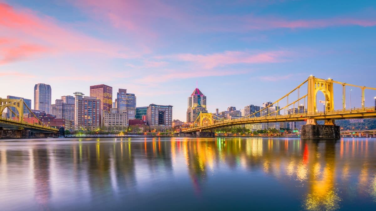 The scene shows two golden bridges crossing a wide river, likely the Allegheny or Monongahela, with the Pittsburgh skyline behind them. The sky features a pink-orange sunset gradient, and the buildings are lit warmly, creating colorful reflections on the water. The overall mood is serene and picturesque, highlighting the city’s iconic bridge architecture and waterfront.