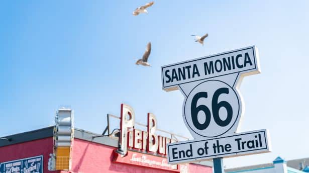 Santa Monica Route 66 "End of the Trail" sign with seagulls flying above and the pier in the background.