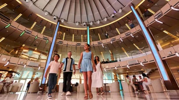Tall, circular glass-domed atrium inside a cruise ship, with guests walking beneath illuminated rings and seating areas.