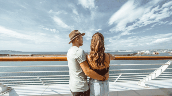 Couple standing together on a cruise ship deck, looking out over the ocean toward a coastal city under a bright blue sky.