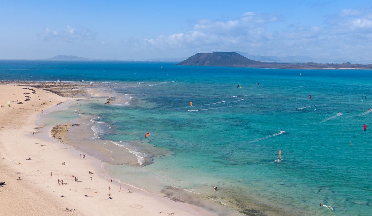 Aerial view of a busy beach with kitesurfers and clear blue waters in Fuerteventura, with distant mountains.