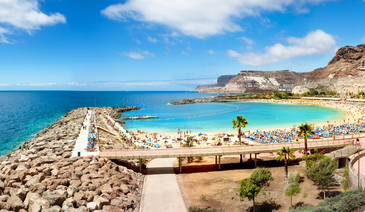 Scenic view of a calm beach in Gran Canaria, featuring golden sands and clear blue waters with tourists enjoying under sun umbrellas.