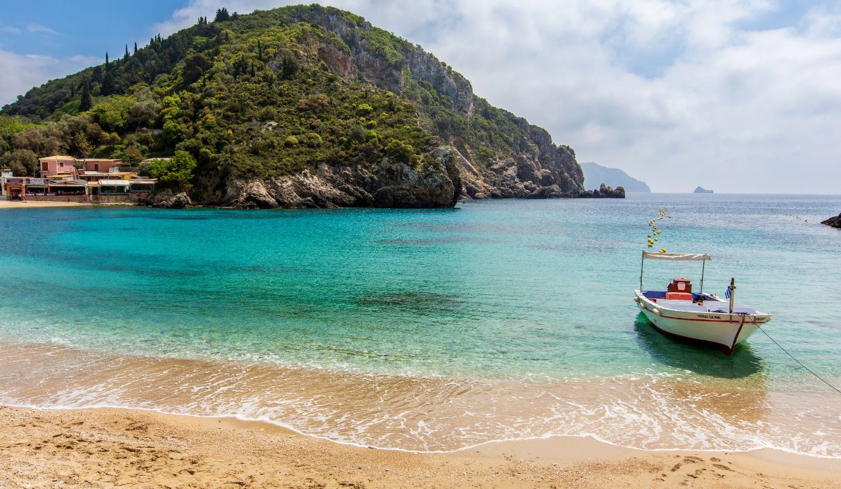 Vibrant beach scene in Corfu with clear aqua waters, white sandy beach, and lush green hills in the background.