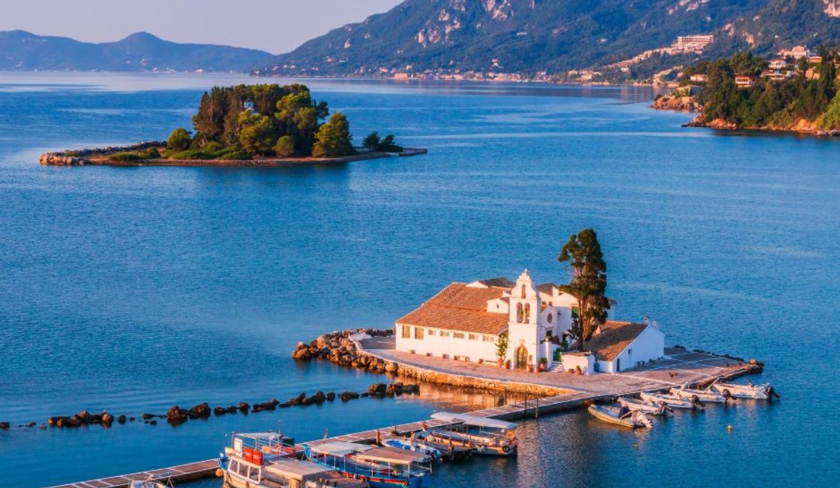 Aerial view of Vlacherna Monastery on a small islet connected by a pier in Corfu, showcasing the surrounding Ionian Sea and lush landscapes.
